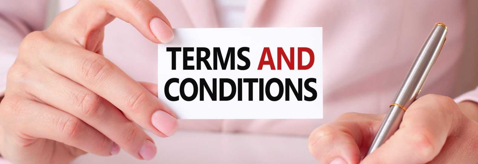 Woman writes in a notebook with a silver pen and hand holding card with text: terms and conditions. Pink background, front view. Business and education concept.